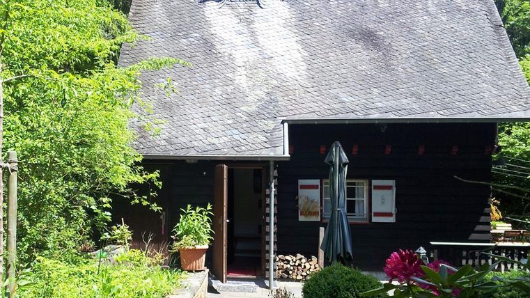 Ein charmantes Holzhaus umgeben von grünem Garten und Bäumen. Die Haustür ist geöffnet und lädt in eine gemütliche Atmosphäre ein.