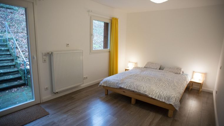 Ein helles Schlafzimmer mit einem Doppelbett und Holzfußboden. Große Fenster bieten Blick auf einen Außenbereich mit Treppen.
