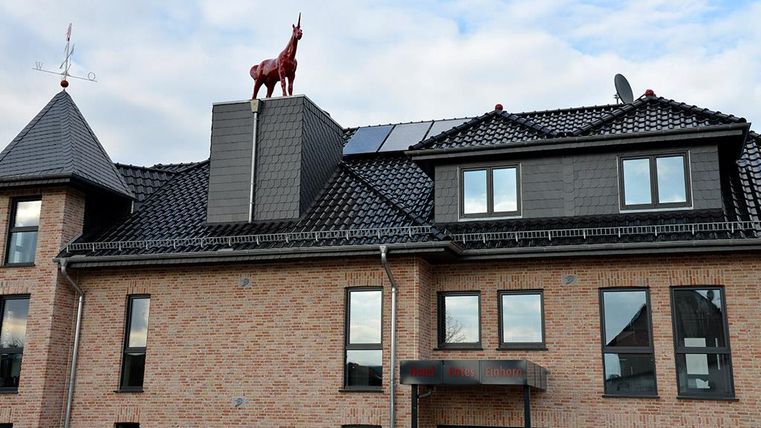 Ein modernes Gebäude mit roten Ziegeln und dunklem Dach. Auf dem Dach steht eine auffällige rote Einhorn-Statue.