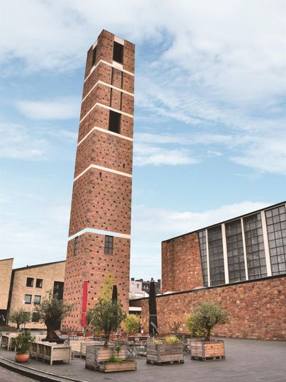 Ein hoher Backsteinturm steht im Zentrum eines Platzes. Um ihn herum sind bepflanzte Sitzbereiche und moderne Gebäude.