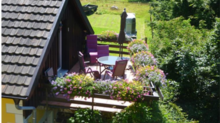 Ein schöner Balkon mit bunten Blumen und gemütlichen Sitzgelegenheiten. Im Hintergrund sieht man eine grüne Wiese und Bäume.