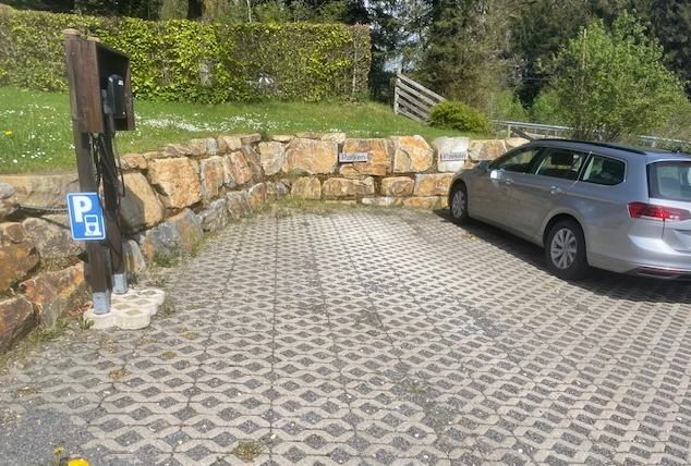 Ein Parkplatz mit einem silbernen Auto und einer Ladestation für Elektrofahrzeuge. Die Umgebung besteht aus Natur und einem gut gepflegten Grünschnitt.