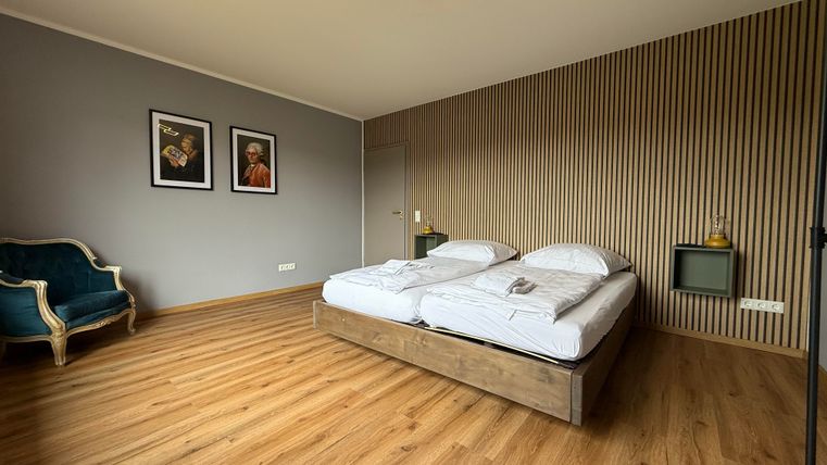 Ein modernes Schlafzimmer mit einem Doppelbett und Holzboden. An der Wand hängen zwei Bilder, und ein eleganter Sessel steht in der Ecke.