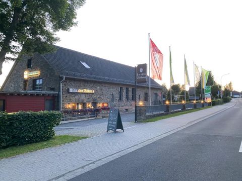 Ein gemütliches Restaurant in einem Steinhaus mit verschiedenen Flaggen vor dem Eingang. Die Umgebung zeigt eine ruhige Straße und grüne Bäume.