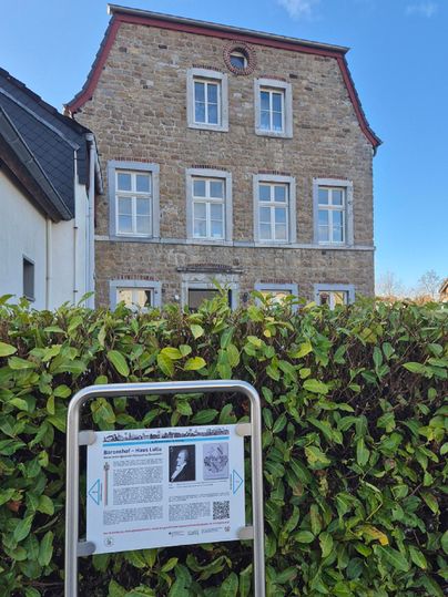 Ein historisches Gebäude mit vielen Fenstern und einem roten Dach. Im Vordergrund befindet sich ein Informationsschild, das mehr über die Geschichte des Hauses erzählt.