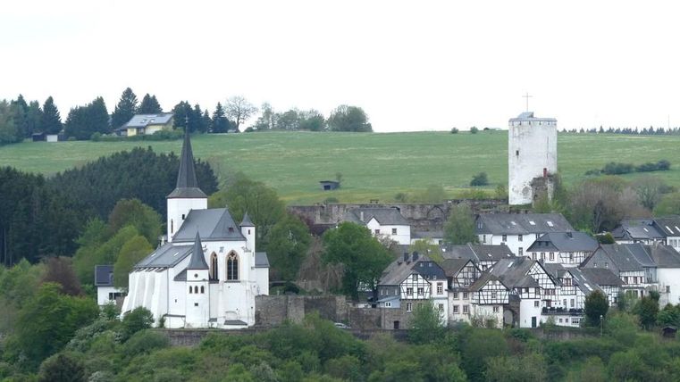 Ein malerisches Dorf mit einer Kirche und traditionellen Häusern. Im Hintergrund sind grüne Hügel und ein Aussichtsturm zu sehen.