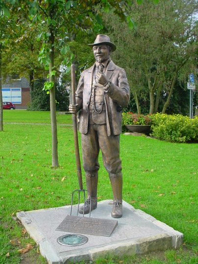 Eine Bronze-Statue eines Mannes mit einem Hut und einer Forke steht auf einem Rasen. Hinter der Statue sind Bäume und Blumenbeete zu sehen.