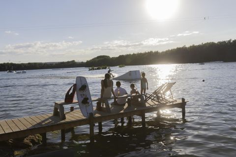 Eine Gruppe von Menschen sitzt auf einem Steg am Wasser. Die Sonne scheint hell und reflektiert auf der Wasseroberfläche.