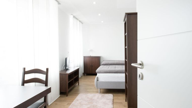 Ein helles, modernes Zimmer mit zwei Betten und einem kleinen Tisch. Die Wände sind in neutralen Farben gehalten, und große Fenster sorgen für viel Tageslicht.