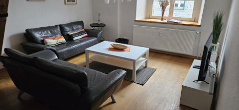 Ein modernes Wohnzimmer mit zwei schwarzen Sofas und einem weißen Couchtisch. Auf dem Tisch liegt eine Dekoration, und ein Fernseher steht auf einem Regal.