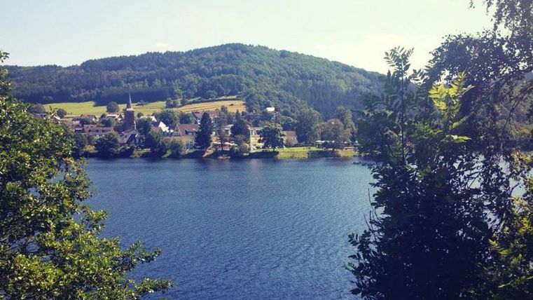 Ein ruhiger See umgeben von sanften Hügeln und Bäumen. Im Hintergrund sind kleine Häuser und eine grüne Landschaft zu sehen.