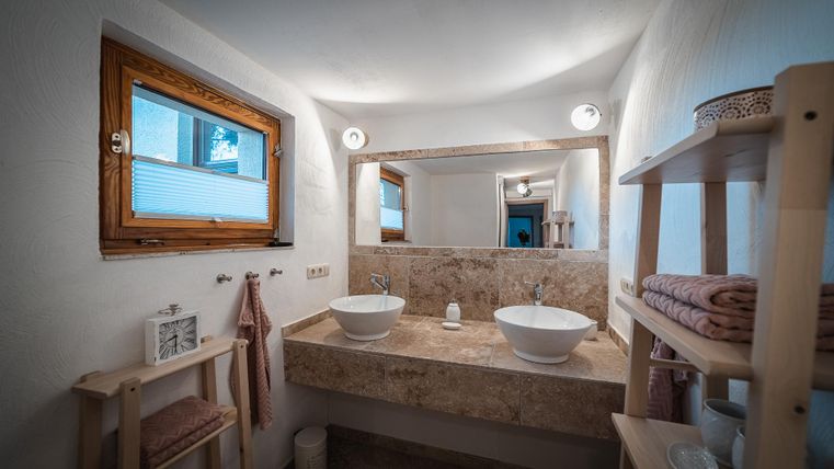 Ein modernes Badezimmer mit zwei Waschbecken und einem großen Spiegel. Die Wände sind hell und es gibt Holzregale mit Handtüchern.