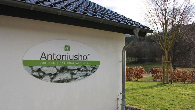 Ein Haus mit einem Schild, das „Antoniushof“ anzeigt. Im Hintergrund sind Bäume und Felder zu sehen.