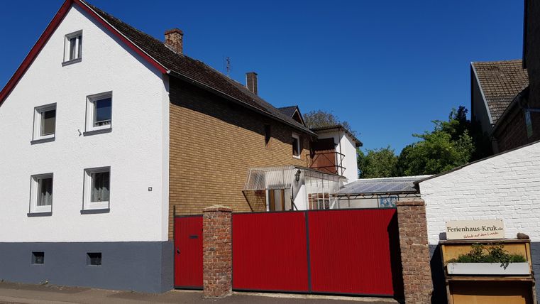 Ein zweigeschossiges Haus mit roten Toren und einer weißen Fassade. Der Himmel ist klar und blau, und es gibt einen kleinen Gartenbereich.
