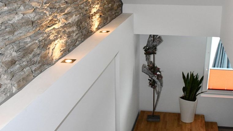 Ein moderner Treppenbereich mit einer Steinwand und stilvollen Lichtern. Neben der Treppe steht eine Pflanze in einem weißen Topf und eine dekorative Skulptur.