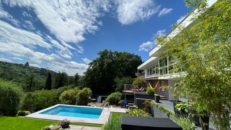 Ein modernes Haus mit einem Schwimmbecken im Garten. Der Himmel ist klar und die Umgebung ist grün und einladend.