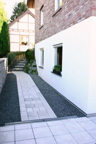 Ein schmaler Weg führt entlang einer weiß gestrichenen Wand eines Hauses, mit einer gepflasterten und kiesbedeckten Fläche. Im Hintergrund sind einige Pflanzen und ein weiteres Gebäude sichtbar.