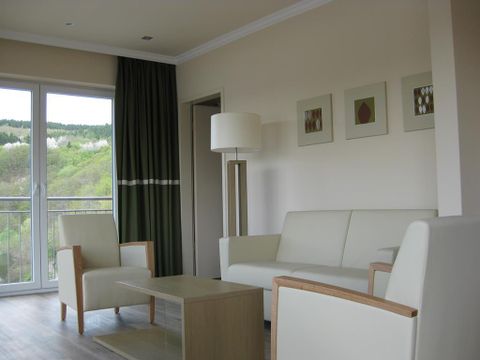 Ein modernes Wohnzimmer mit einem weißen Sofa und Holzstühlen. Große Fenster bieten einen Blick auf die grüne Landschaft.