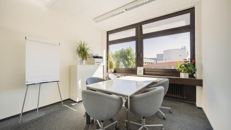 Ein modernes Besprechungszimmer mit einem Tisch und fünf Stühlen. Große Fenster bieten einen Blick auf einen grünen Außenbereich.