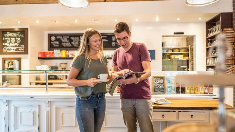 Ein Paar steht in einem Café und betrachtet eine Speisekarte. Sie halten Tassen in der Hand und lächeln.