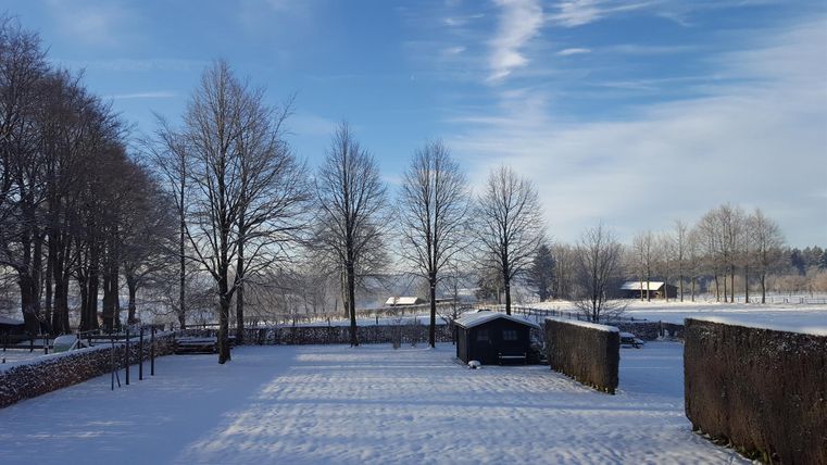 Eine winterliche Landschaft mit schneebedeckten Bäumen und klarem Himmel. Im Hintergrund sind kleine Hütten und Felder sichtbar.