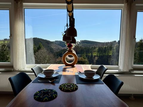 Ein gemütlicher Essbereich mit einer Holztafel und zwei Schalen. Durch das große Fenster sieht man eine malerische Landschaft mit Hügeln und Bäumen unter einem klaren blauen Himmel.