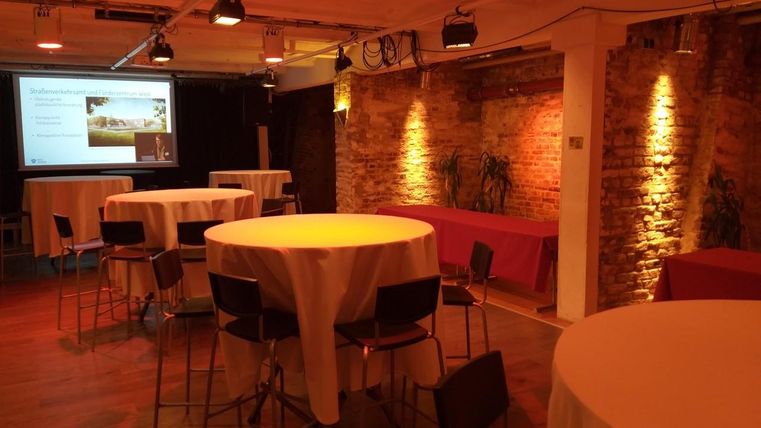 Ein moderner Veranstaltungsraum mit runden Stehtischen und warmem Licht. Im Hintergrund sind eine Präsentationsleinwand und einige Pflanzen zu sehen.