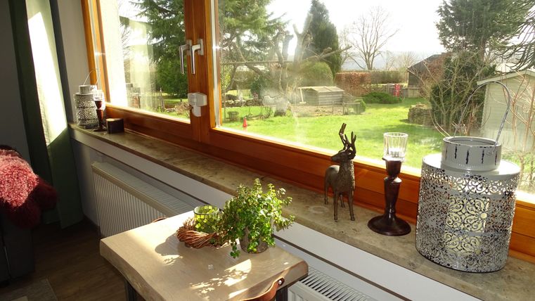 Ein gemütlicher Raum mit einem großen Fenster, das einen Blick auf einen grünen Garten bietet. Auf der Fensterbank stehen Pflanzen und dekorative Gegenstände.