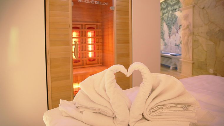 Ein entspannendes Wellnesszimmer mit zwei handtuchförmigen Schwänen, die ein Herz bilden. Im Hintergrund steht eine Infrarotsauna mit warmem Licht.