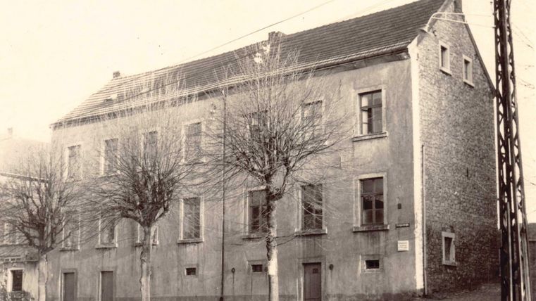 Ein altes Gebäude mit einer einfachen Fassade und mehreren Fenstern. Vor dem Gebäude stehen kahle Bäume und eine Straßenlaterne.