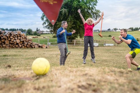 Drei Personen haben Spaß auf einer Wiese. Sie feiern und springen vor Freude, während ein gelber Ball im Vordergrund liegt.