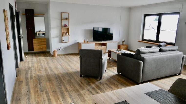 Eine moderne Wohnziemmmer mit einem grauen Sofa und einem Holztisch. Der Raum ist hell mit großen Fenstern und einem Fernseher an der Wand.