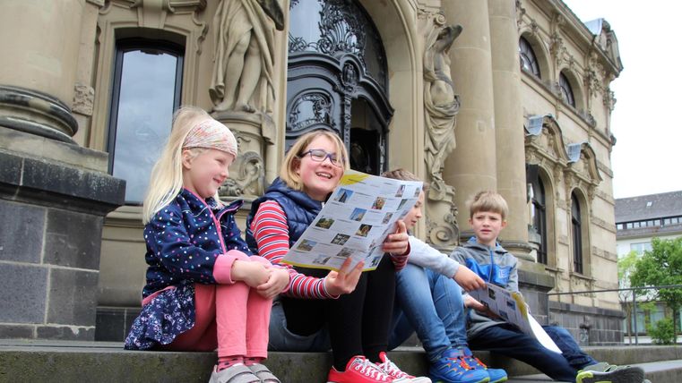 Drei Kinder sitzen auf der Treppe eines historischen Gebäudes. Sie schauen auf eine Karte und scheinen einen Ausflug zu planen.