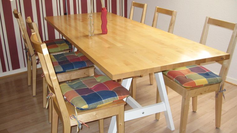 Ein einfacher Esstisch aus Holz mit bunten Stühlen. Im Hintergrund sind gestreifte Wände zu sehen.