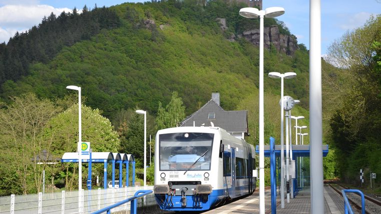 Ein Zug wartet an einer Haltestelle, umgeben von grünen Hügeln. Die Sonne scheint und es ist ein klarer Tag.