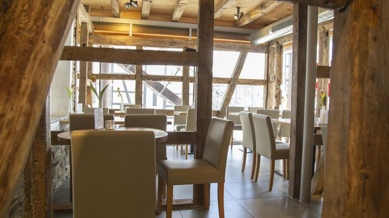 Ein gemütliches Restaurant mit Holzbalken und eleganten Tischen. Die große Fensterfront sorgt für viel Licht und eine einladende Atmosphäre.
