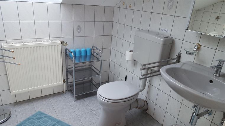 Ein Badezimmer mit weißen Fliesen, einem Waschbecken und einer Toilette. Es gibt auch einen Handtuchhalter und einen kleinen Stauraum für Toilettenartikel.
