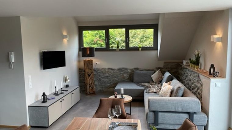 Ein modernes Wohnzimmer mit einer gemütlichen Couch und einem Holztisch. Große Fenster lassen viel Licht herein und bieten einen Blick auf die Natur draußen.