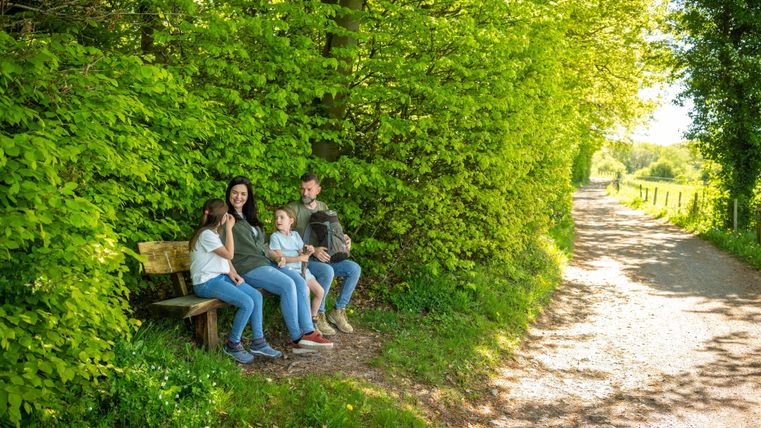 Eine Familie sitzt auf einer Bank neben einem schattigen Weg. Der Weg ist von üppigem Grün und Bäumen umgeben.