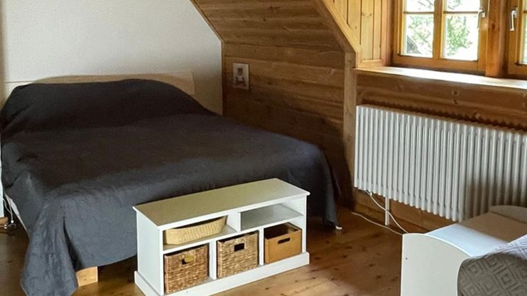 Ein gemütliches Schlafzimmer mit einem großen Bett und Holzwänden. Es gibt ein Regal mit Aufbewahrungskörben und große Fenster für viel Tageslicht.