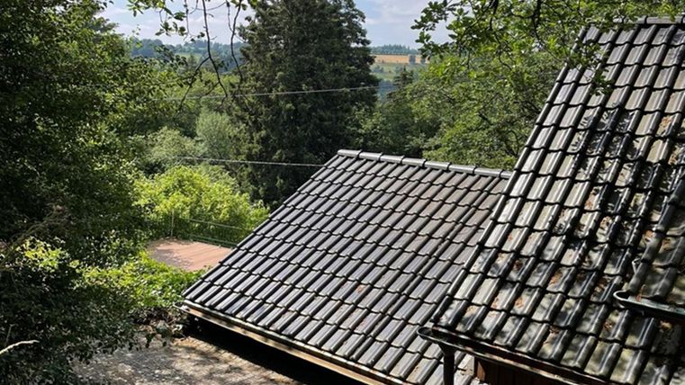 Ein Blick auf die Dächer eines Hauses, umgeben von grünen Bäumen und einem ruhigen Landschaftshorizont. Der Himmel ist teilweise bewölkt, was eine entspannte Atmosphäre schafft.