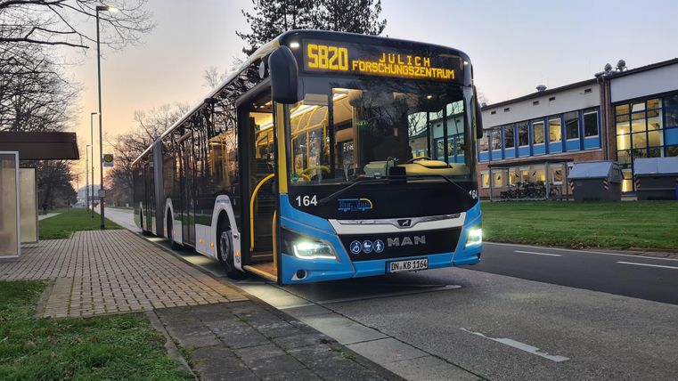 Ein moderner Bus hält an einer Haltestelle. Im Hintergrund sind Gebäude und Bäume zu sehen.
