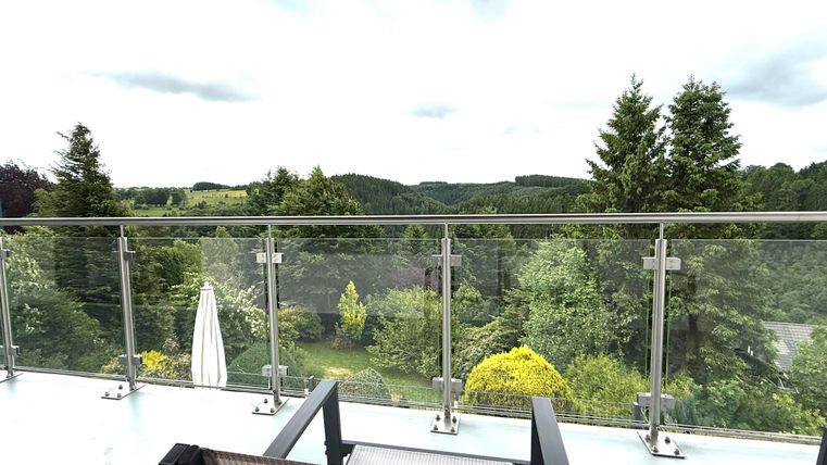 Ein Balkon mit Blick auf eine grüne Landschaft. Die Umgebung ist geprägt von Bäumen und Hügeln unter einem bewölkten Himmel.