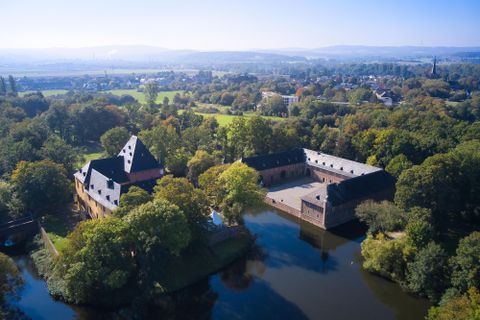 Ein malerisches Schloss umgeben von Bäumen und einem ruhigen Wasser. Im Hintergrund sind sanfte Hügel und eine klare Himmelssicht zu sehen.