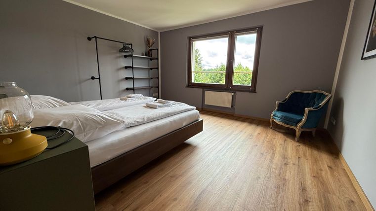 Ein modernes Schlafzimmer mit einem Doppelbett und großem Fenster. Der Raum hat Holzboden und eine gemütliche Sitzgelegenheit in der Ecke.