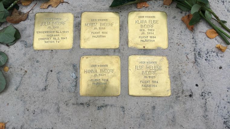Sechs Gedenktafeln aus Messing liegen auf dem Boden, umgeben von Rosenblättern. Jede Tafel trägt den Namen und die biografischen Details von Personen.