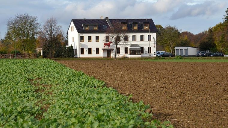 Ein weißes Haus steht neben einem Ackerfeld. Auf der linken Seite wächst frisches Grün, während der Boden auf der rechten Seite bearbeitet ist.
