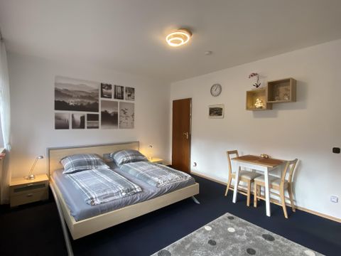 Ein modernes Schlafzimmer mit einem Doppelbett und Holzschränken. Ein kleiner Tisch mit Stühlen und dekorative Bilder an der Wand verleihen dem Raum Gemütlichkeit.