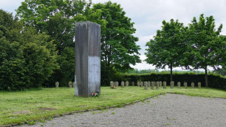 Ein Denkmal aus Stein steht in einer grünen Wiese, umgeben von Bäumen. Rund um das Denkmal sind kleine Steine aufgestellt.
