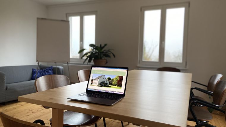 Ein moderner Besprechungsraum mit einem großen Tisch aus Holz. Auf dem Tisch steht ein Laptop, daneben eine Pflanze und ein Sofa.